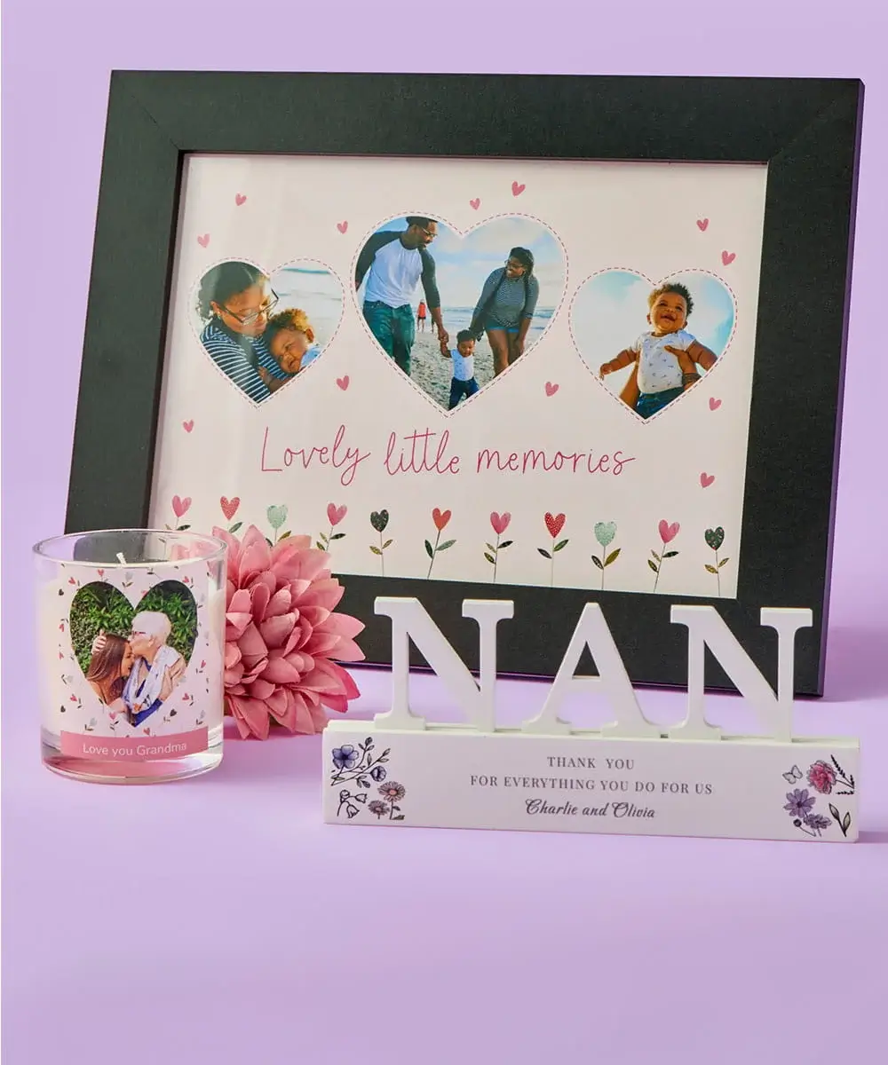 A heart‑themed photo frame with three family photos sits behind a personalised candle and a decorative NAN ornament, all arranged with a pink flower on a pastel purple background.